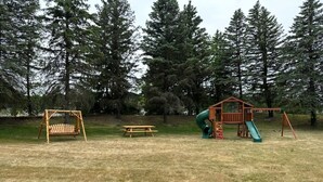 Un parque infantil con columpios, un tobogán y una pequeña estructura de madera en un área verde con árboles al fondo.