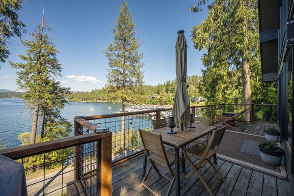 A wooden deck with a table and chairs, overlooking a lake with trees and a dock.