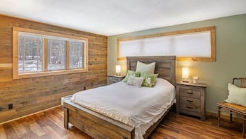 A bedroom with a wooden bed, two bedside tables, a nightstand, and a window with a view of trees.