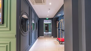 Un couloir d’hôtel avec un mur végétal, un miroir, un chariot à bagages rouge et un plafond doté d’un éclairage encastré.
