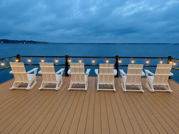 Une terrasse avec des chaises blanches et des guirlandes lumineuses, donnant sur une étendue d’eau.