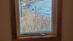 A room with a window overlooking a rocky shoreline with trees and a house. - Superior Reflections  Cabin #2 on Lake Superior (Schroeder)