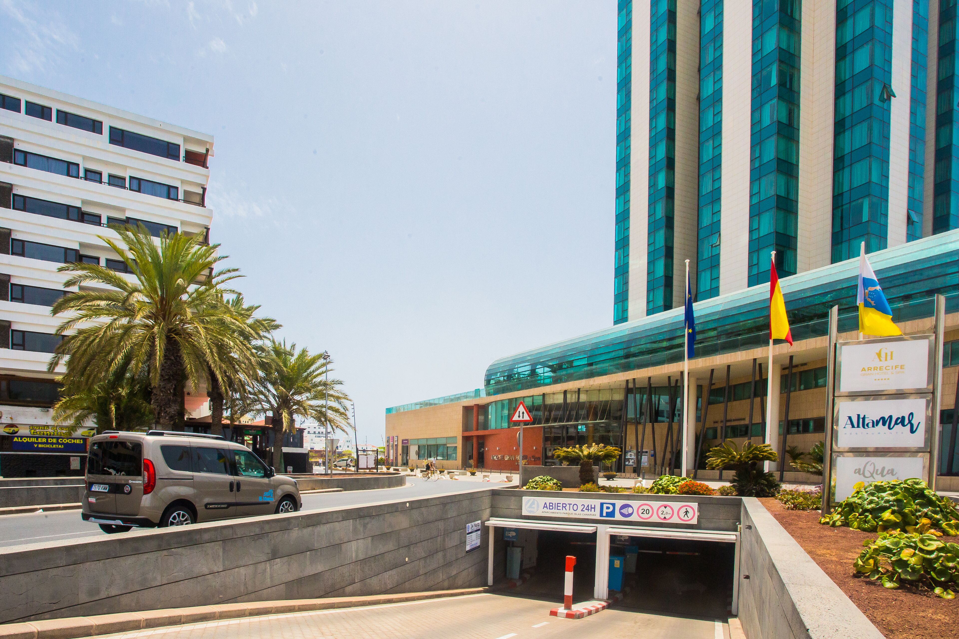 Un edificio moderno con fachada de vidrio, palmeras y una furgoneta estacionada al frente.