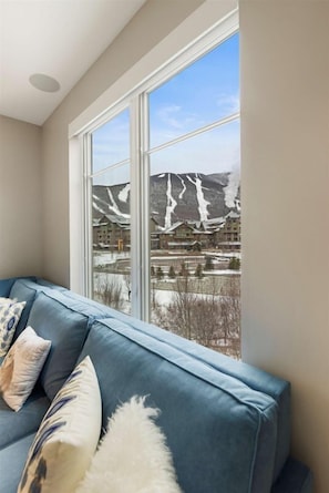 A room with a large window, a blue sofa, and a view of a mountain and lodge.