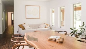 A modern living room with a wooden dining table, a sofa set, and large windows.