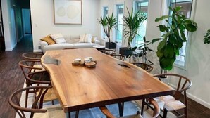 A spacious dining area with a large wooden table and wicker chairs, a white sofa, and several potted plants.