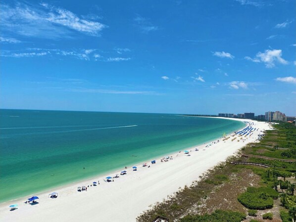 A beach with a clear sky, a sandy shore, and a few buildings in the distance.