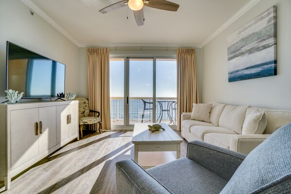 A modern living room with a flat-screen TV, a sofa, a coffee table, and a balcony with a view of the sea.