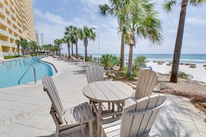 A poolside area with chairs and tables, a pool, palm trees, and a building in the background.