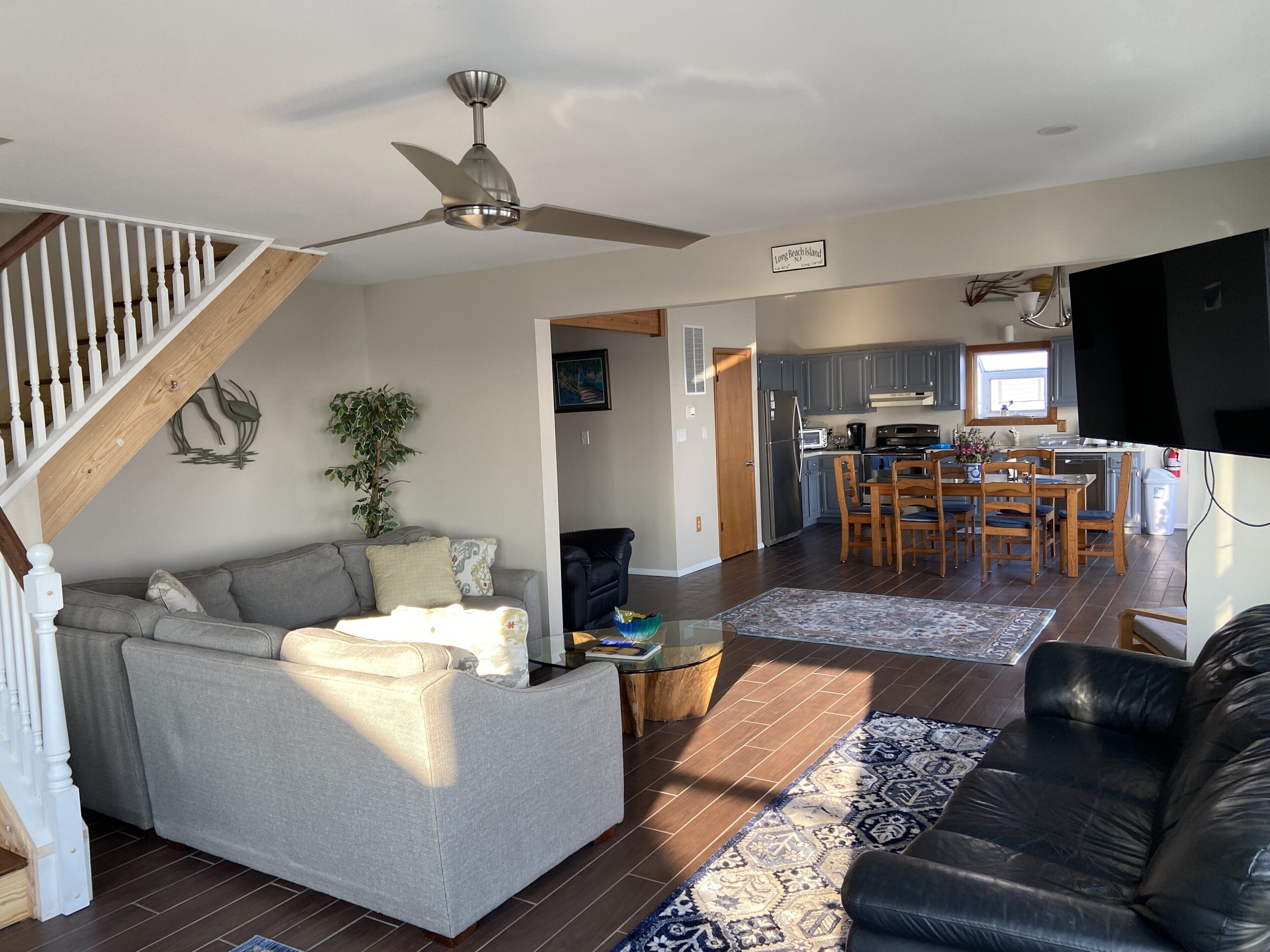 A spacious living area with a gray sofa, a black leather couch, and a dining table with chairs. A ceiling fan is visible, and a kitchen area is in the background.