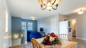 Un comedor con una mesa preparada para dos personas, un sillón tipo bean bag azul, un televisor al fondo y una cocina visible a través de una puerta abierta.