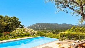 A pool with lounge chairs and a table, surrounded by greenery and mountains in the background.