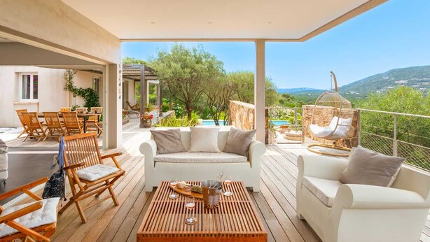 A spacious outdoor patio with wooden furniture, a white sofa, and a hanging chair overlooking a pool and greenery.