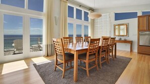 Uma área de refeições com mesa e cadeiras de madeira, janelas amplas com vista para o oceano, cozinha com armários de madeira e uma luminária pendente.