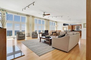 A spacious living room with a large sofa, armchairs, and a coffee table. There are large windows offering a view of the ocean.