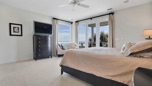 Quarto com cama, ventoinha no teto, televisão, cômoda e vista para o exterior.