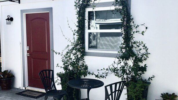 Un edificio blanco con puerta roja, una ventana y un pequeño patio con dos sillas y una mesa.