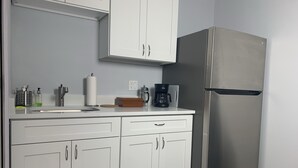 Una cocina con armarios blancos, un refrigerador de acero inoxidable, una cafetera y un lavamanos con distintos artículos de tocador.
