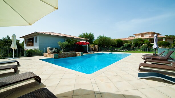 A swimming pool with lounge chairs and umbrellas, surrounded by a fence and greenery. - Beautiful apartment in Residence with swimming pool, garden and terrace (Sainte-Lucie de Porto-Vecchio)