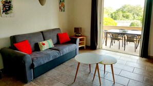 A living room with a grey sofa, red and multicolored pillows, a small table with a lamp, and a view of an outdoor patio with a table and chairs. - Beautiful apartment in Residence with swimming pool, garden and terrace (Sainte-Lucie de Porto-Vecchio)