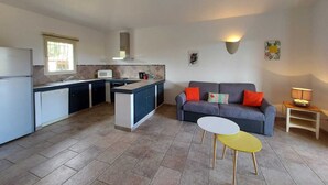A compact kitchen with a built-in microwave, a gas stove, and a sink. There is a grey sofa with orange cushions and a small yellow table. - Beautiful apartment in Residence with swimming pool, garden and terrace (Sainte-Lucie de Porto-Vecchio)