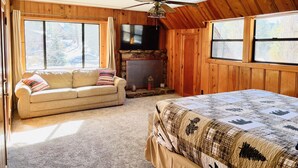 Une pièce chaleureuse avec un plafond en bois, un ventilateur de plafond, un grand lit recouvert d’une courtepointe, un canapé agrémenté d’un coussin à motifs, une cheminée et de grandes fenêtres.