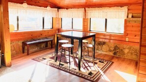 L’intérieur chaleureux d’un chalet avec des murs en bois, une cheminée en pierre, une table en bois entourée de tabourets et de grandes fenêtres ornées de rideaux en dentelle.