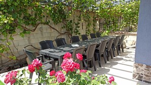 An outdoor dining area with a long table and chairs, surrounded by greenery and a stone wall. - Fabulous holiday home with sea view for the whole family! (Balchik)