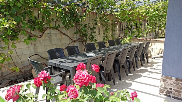 An outdoor dining area with a long table and chairs, surrounded by greenery and a stone wall.