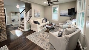 Un salon spacieux avec un canapé blanc, un fauteuil à rayures et une table basse. On peut voir un ventilateur de plafond, et un escalier mène à un autre niveau.
