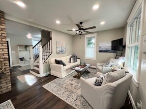 Un salon spacieux avec un canapé blanc, un fauteuil à rayures et une table basse. On peut voir un ventilateur de plafond, et un escalier mène à un autre niveau.
