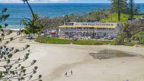 A beachside hotel with a view of the ocean, a parking area, and a building with a sign reading "FROM WHERE YOU'D RATHER BE".