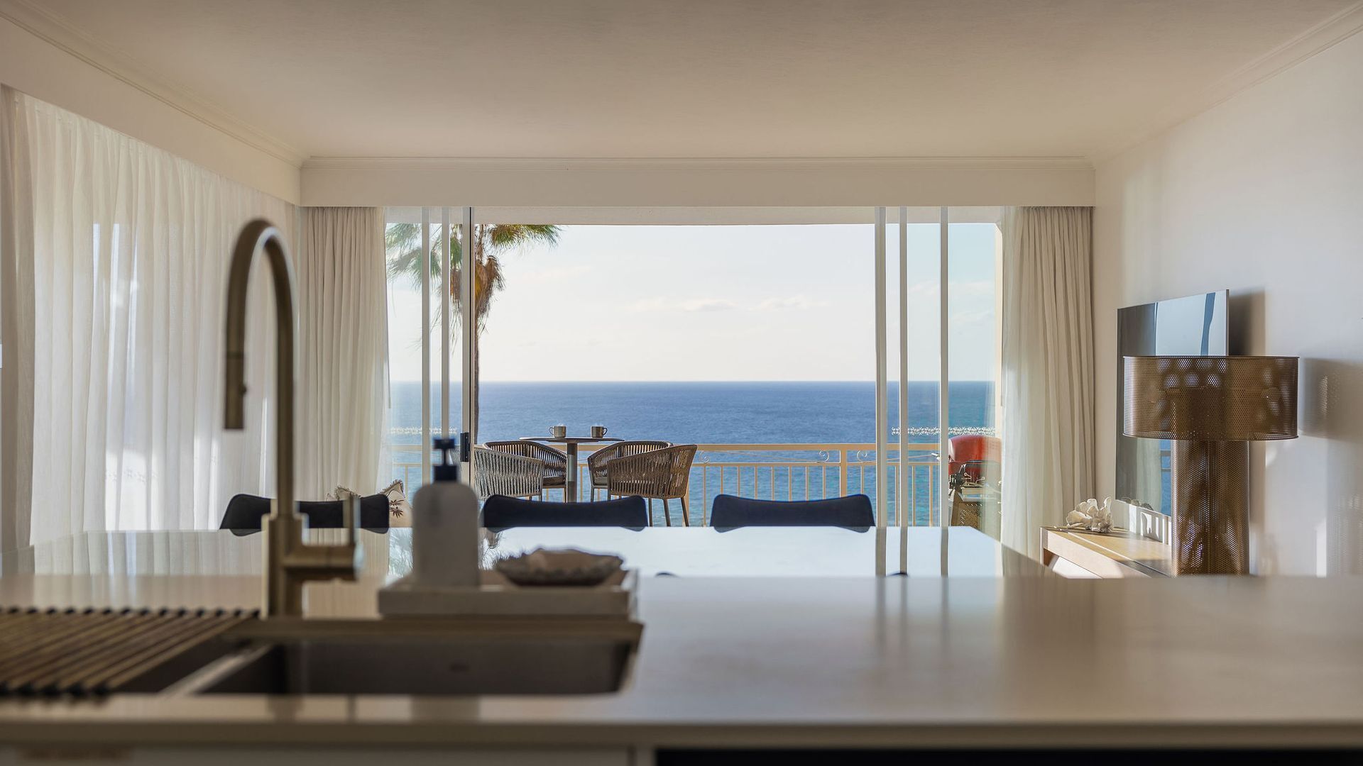 Habitación de hotel con balcón que da al mar, zona de comedor con sillas y mesa, y televisor montado en la pared.