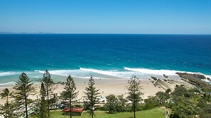 A coastal view with a beach, clear blue sea, and a distant city skyline.