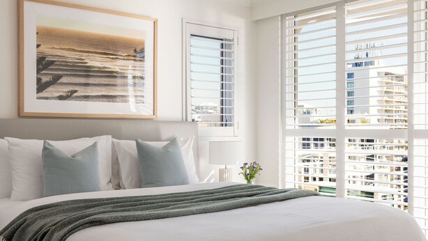 A hotel room with a bed, pillows, a framed picture, a window with blinds, and a view of buildings. - Orion 6 - Beachfront Rainbow Bay - All Linen Supplied - Min. 3 Night Stays! (Rainbow Bay)