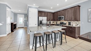 A spacious kitchen with dark wooden cabinets, a large island with bar stools, and a microwave. - Grand Traverse Paradise | 8-Bedroom Villa w/ Private Pool at Reunion Resort (Kissimmee)