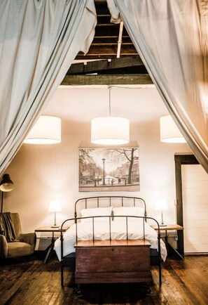 A bedroom with a metal bed frame, two bedside tables with lamps, a wooden floor, and a large window with curtains. - THE STARR LOFT- Earlham, IUEast, Historic District (Richmond)