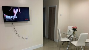 A modern dining area with a glass table and white chairs, a television mounted on the wall, and a closed door in the background. - Everything is pretty much walking distance which is odd for Miami  (Hialeah)
