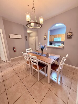 A dining area with a wooden table, white chairs, a chandelier, and a kitchen visible through an archway. - Windsor Wonderland!  Your Disney Getaway! Water slides, pool, arcade and more! (Kissimmee)