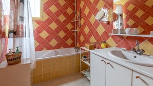 A bathroom with a bathtub, a sink, and a shower. There is a window with a curtain, and the walls are tiled with a diamond pattern. - Villa Bagatelle, et son jardin - Welkeys (Montpellier)