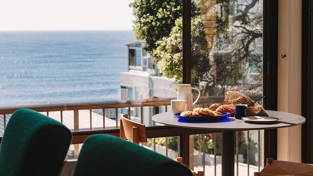 Une table ronde avec une carafe, une tasse et des pâtisseries, le tout placé sur un balcon donnant sur la mer.