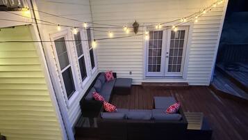 Een overdekt terras met een donkerbank en kussens, een houten terras en lichtslingers.