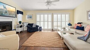 Ein Wohnzimmer mit Kamin, Deckenventilator, einer Couch, einem Couchtisch und einem großen Fenster mit Jalousien.