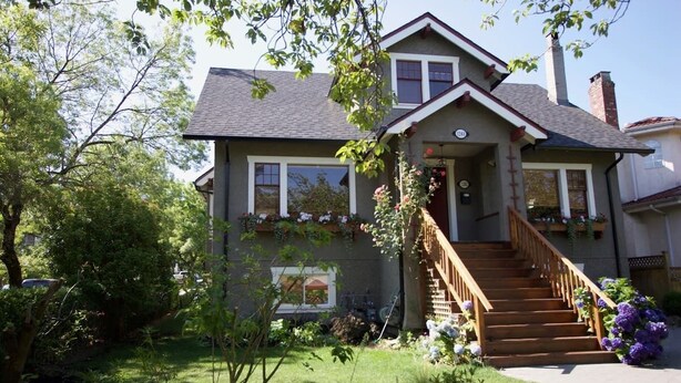 A two-story house with a front porch, wooden stairs, and a garden with flowers and shrubs.