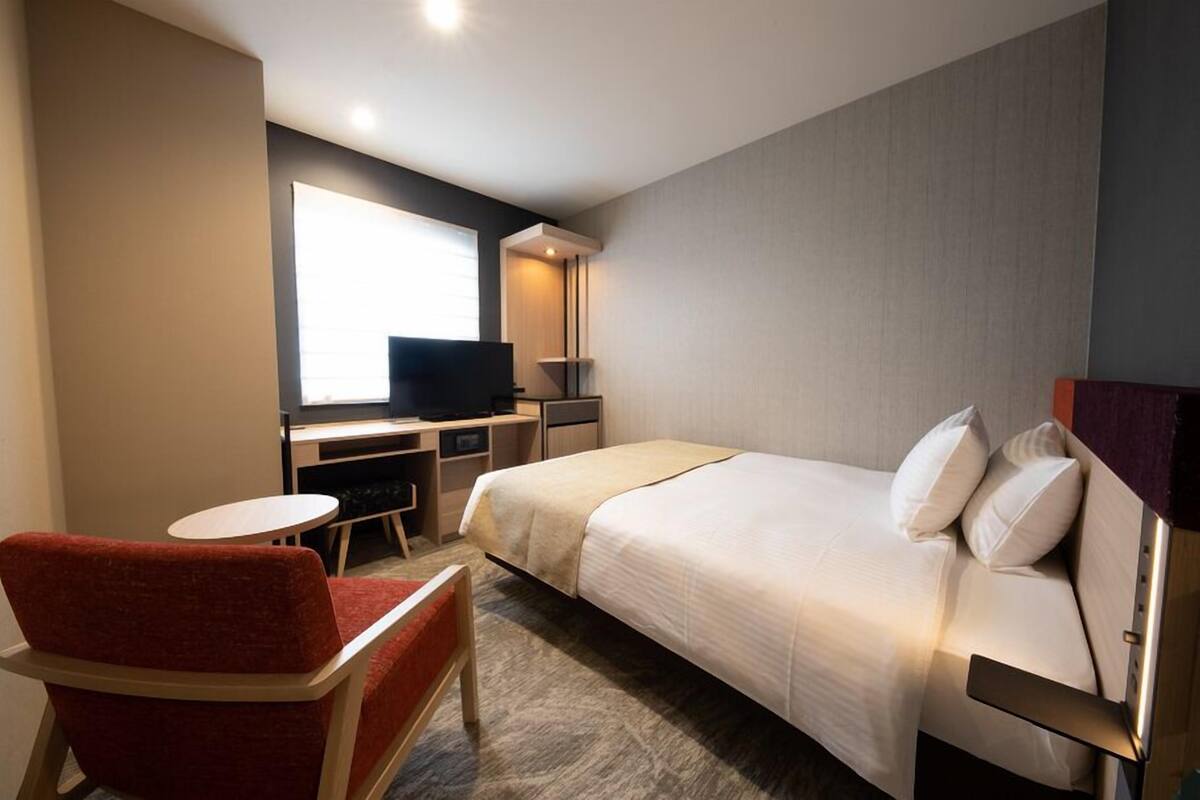 A hotel room with a bed, a desk with a television, a chair, and a small table.