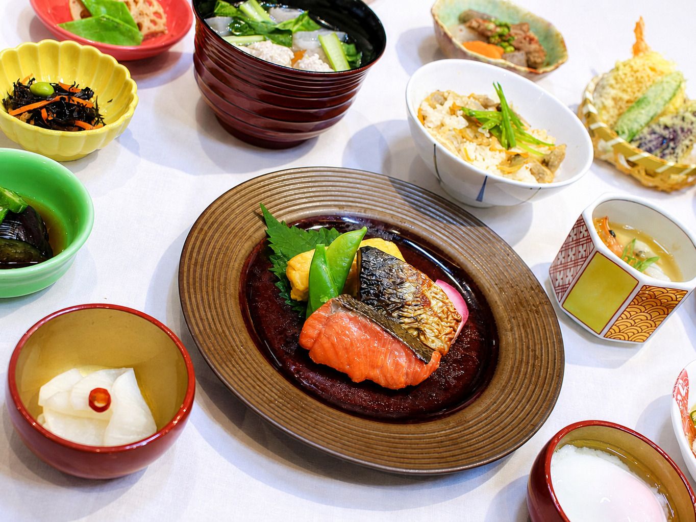 Variedad de platos sobre una mesa, incluyendo un plato central con pescado y verduras, rodeado de recipientes más pequeños con diferentes alimentos.