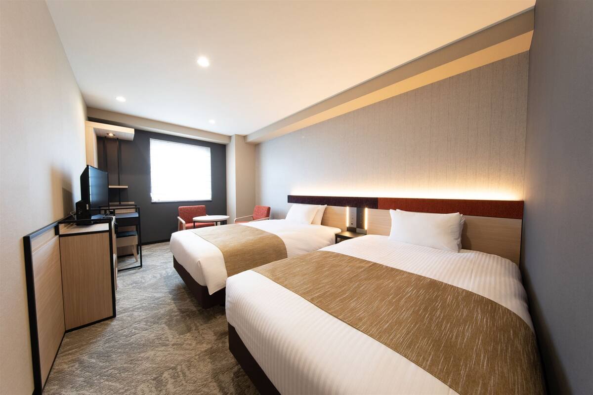 A hotel room with two beds, a desk, a chair, a TV, and a window.