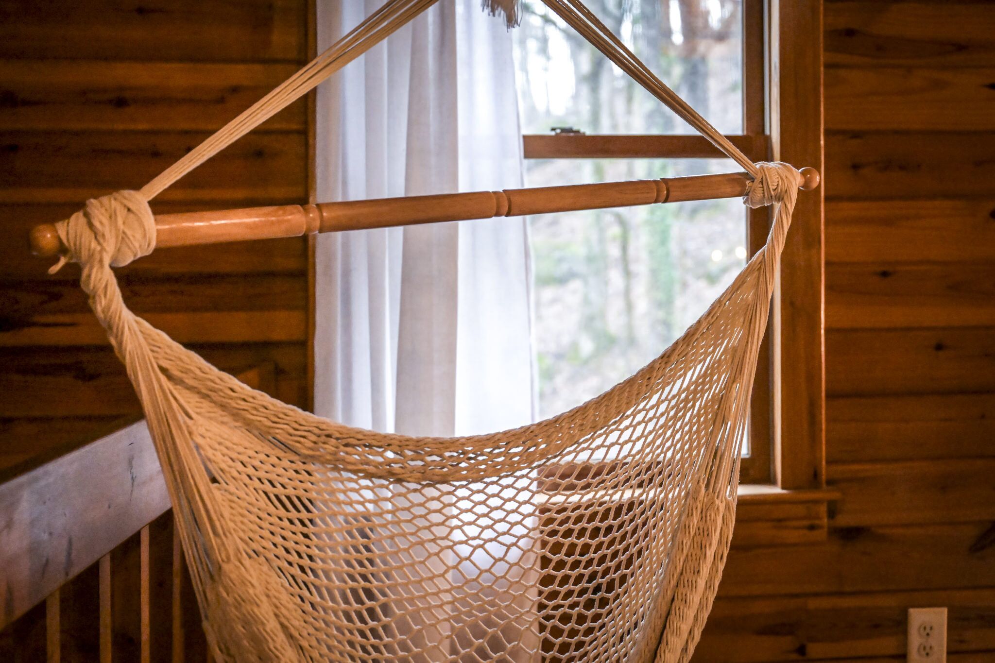 Una hamaca atada a una viga de madera en una cabaña con paredes y ventana de madera.