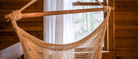 Una hamaca atada a una viga de madera en una cabaña con paredes y ventana de madera.