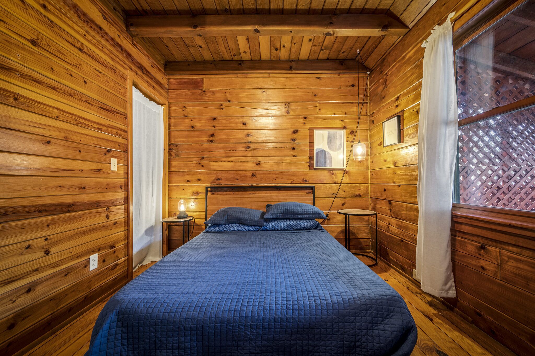 A wooden cabin room with a bed, a small table, and a window with curtains.